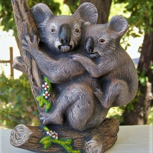 May include: A ceramic figurine of two koalas hugging on a tree branch. The koalas are painted in shades of brown and gray, with white accents. The tree branch is painted brown with green leaves and white flowers.