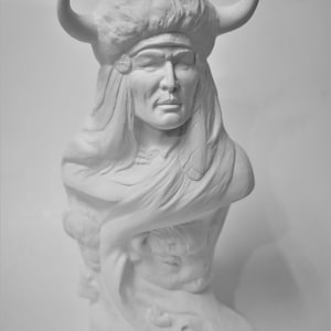 May include: White ceramic bust of a Native American man wearing a headdress with horns. The bust is detailed with feathers and a beaded necklace.