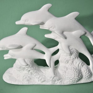May include: White ceramic sculpture of four dolphins leaping out of the water. The dolphins are arranged in a playful formation, with their tails and fins extended. The sculpture is set against a textured background that resembles waves.