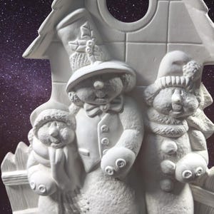May include: White ceramic figurine of three snowmen standing in front of a house. The snowman in the middle is wearing a hat and a scarf. The snowman on the left is wearing a scarf. The snowman on the right is wearing a hat.