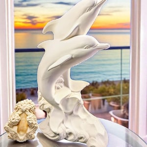 May include: A white dolphin sculpture with two dolphins leaping from a wave base. The sculpture is on a glass table, with a seashell and a small glass bowl nearby. The background shows a sunset over the ocean.
