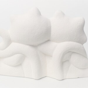 May include: White ceramic figurine of two cats cuddling together. The cats are sitting side by side with their tails intertwined.