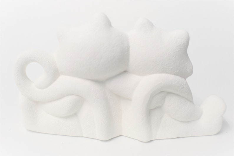 May include: White ceramic figurine of two cats cuddling together. The cats are sitting side by side with their tails intertwined.