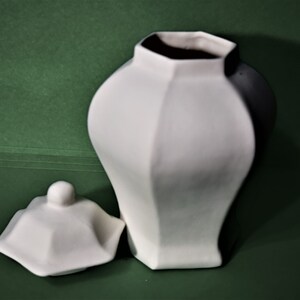 Peut inclure: Un vase en c&eacute;ramique blanc avec un design g&eacute;om&eacute;trique et un couvercle assorti. Le vase a un corps bulbeux et un col hexagonal &agrave; facettes. Le couvercle est fa&ccedil;onn&eacute; pour s'adapter au vase, avec un petit bouton sur le dessus. Le fond est vert uni.