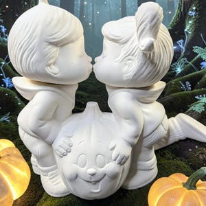 May include: A white ceramic figurine depicting two children leaning over a carved pumpkin to kiss. The children have detailed hair and clothing, while the pumpkin features a smiling face. The sculpture is matte white and stands as a single piece.