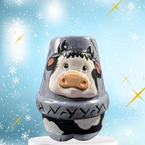 May include: A gray ceramic planter shaped like a cow with black and white spots. The planter has a black nose, black eyes, and black horns. The planter is sitting on a white pedestal.