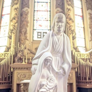 May include: A white religious statue depicting the Holy Family: Joseph, Mary, and baby Jesus. The statue is in a church setting with stained glass windows and ornate architectural details in the background. The figures are draped in flowing robes.