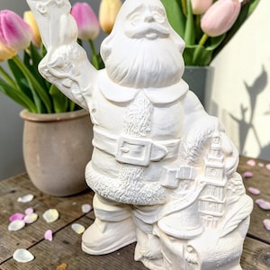 May include: A white ceramic Santa Claus figurine holding a letter and a bag. The Santa figure has a long white beard, a belt, and a fur-trimmed coat. The bag has a scroll and a building design. The background includes tulips.