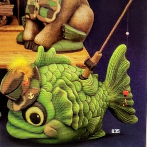May include: A green ceramic fish figurine with a large eye, wearing a camouflage hat with an anchor emblem. The fish has a fishing rod sticking out of its back, with a red bobber on the line. The number 835 is visible in the bottom right corner of the image.