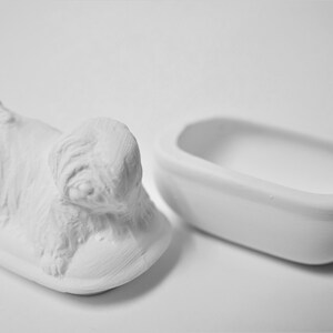 K&ouml;nnte beinhalten: Wei&szlig;e Keramik-Schmuckschatulle in Form eines liegenden Hundes. Die Schachtel ist ge&ouml;ffnet und der Deckel ist separat. Der Deckel ist in Form einer kleinen Badewanne.