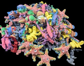 May include: A pile of colorful, articulated animal puzzles in various shapes, including dogs, lizards, and starfish. The puzzles are primarily pastel shades of pink, blue, yellow, and green, with some gray and black accents. The background is solid black.