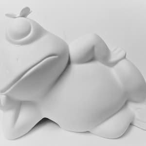 May include: A white ceramic frog figurine in a relaxed pose, with its arms crossed and a small butterfly perched on its head. The frog is lying down, with its eyes closed, and has a whimsical, cartoonish design.