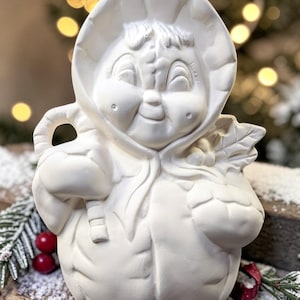 May include: A matte white ceramic figurine of a child wearing a hooded winter coat. The figure has a smiling expression, holds a candy cane in one hand, and carries a cluster of holly leaves and berries in the other, featuring a smooth finish.
