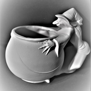 May include: White ceramic cauldron with a witch figurine holding onto the side. The witch is wearing a pointed hat and has a long, flowing robe.