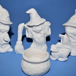 May include: A collection of unpainted ceramic Halloween figurines. The set includes three witch figures wearing pointed hats, a cauldron, a small dog, and a broom. The figures are all white and ready to be painted.