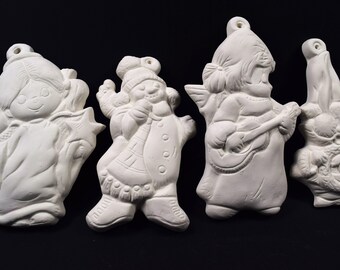 May include: Four unpainted ceramic Christmas ornaments in various shapes. The ornaments include a girl holding a star, a snowman with a sack, an angel playing a guitar, and Santa Claus with a wreath. Each ornament has a hole for hanging.
