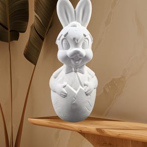 May include: White ceramic bunny rabbit figurine emerging from a cracked egg. The bunny has a bow tie and is holding the egg with its paws.