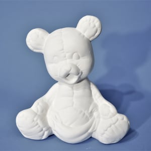 May include: White ceramic teddy bear figurine sitting with its legs crossed. The bear has a stitched-like texture and a simple, friendly face.