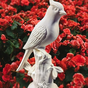 May include: A white ceramic bird statue perched on a tree stump, set against a backdrop of vibrant red flowers. The bird has detailed feather and eye features, and the stump has carved bark and floral accents.