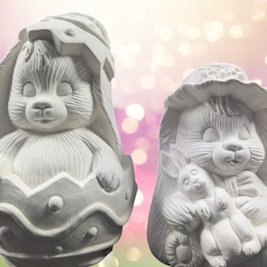 May include: Two white ceramic bunny figurines. One bunny is sitting inside an egg-shaped shell. The other bunny is holding a smaller bunny figurine.