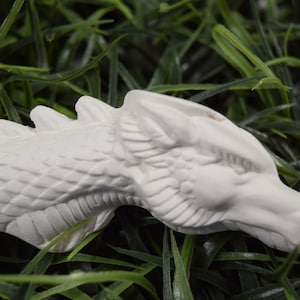 May include: White ceramic dragon head figurine with detailed scales and a textured surface. The figurine is lying on a bed of green grass.