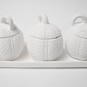 May include: Three white ceramic pumpkin-shaped containers with lids, arranged on a white rectangular tray. The containers have a textured, leaf-like design.