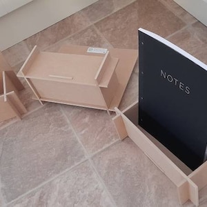 May include: A collection of unfinished wooden desk organizers and a notebook. The notebook has a black cover with the word "NOTES" in white. The organizers are various shapes and sizes, including a box and a stand.