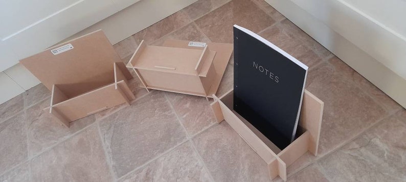 May include: A collection of unfinished wooden desk organizers and a notebook. The notebook has a black cover with the word "NOTES" in white. The organizers are various shapes and sizes, including a box and a stand.