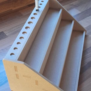 May include: A light brown wooden desk organiser with multiple compartments and a row of circular holes for pens. A blue and white pen is placed in one of the holes. The organiser has a tiered design.