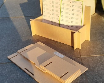 May include: A light brown wooden box with a slot for holding items, and a stack of unassembled wooden pieces. The box holds several sheets of printed labels. The pieces have slots for assembly. The box is on a grey surface.
