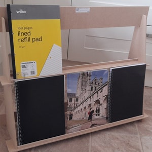 May include: A light-colored wooden desktop organizer holding a Wilko 160 pages lined refill pad, two black spiral-bound notebooks, and a notebook featuring a photograph of a stone cathedral and a dancer in a black outfit on the cover.