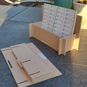 May include: A light brown, unfinished wooden box with a stack of matching wooden pieces. The box holds a stack of printed sheets with a logo. The wooden pieces have slots and tabs for assembly. The box and pieces are on a tiled surface.