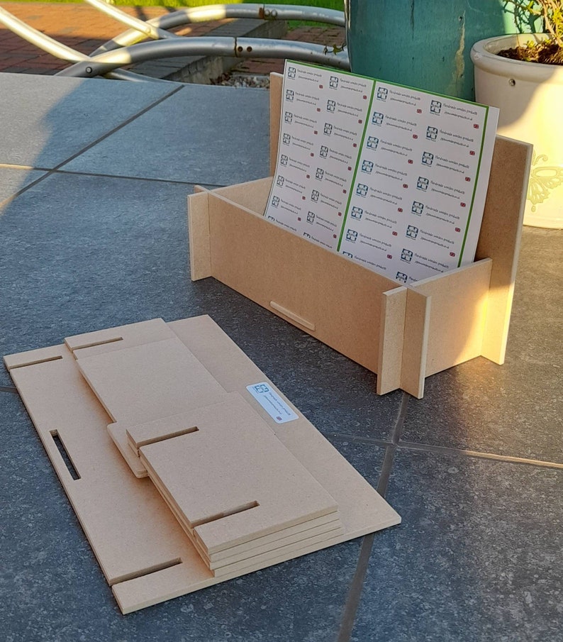 May include: A light brown, unfinished wooden box with a stack of matching wooden pieces. The box holds a stack of printed sheets with a logo. The wooden pieces have slots and tabs for assembly. The box and pieces are on a tiled surface.