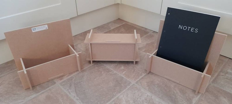 May include: Three unfinished wooden organizers. One holds a black notebook with "NOTES" printed on it. The other two are open boxes. The organizers are made of light-colored wood and are designed to hold items.