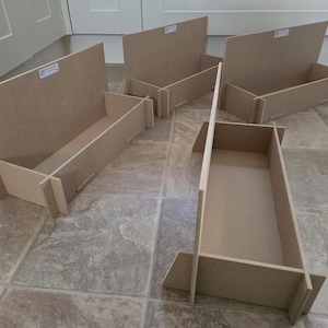 May include: Five unfinished, light brown wooden storage boxes. Each box has a rectangular base and angled sides, with a back panel. The boxes are designed to be assembled and used for organizing items. They are placed on a tiled floor.