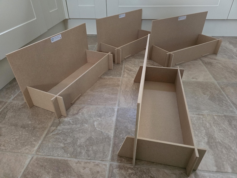 May include: Five unfinished, light brown wooden storage boxes. Each box has a rectangular base and angled sides, with a back panel. The boxes are designed to be assembled and used for organizing items. They are placed on a tiled floor.