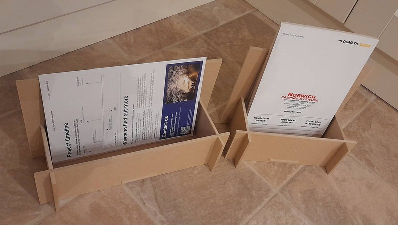 May include: Two unfinished wooden display stands, each holding printed materials. One stand displays a document with a timeline and contact information. The other features a brochure for Norwich Camping & Leisure.