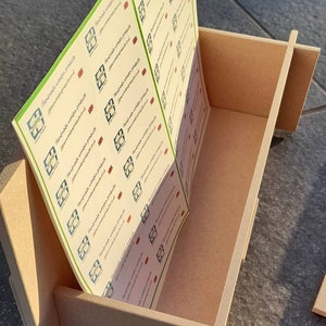 May include: A light brown wooden box with a slotted design, holding a stack of printed labels. The labels have a green border and feature a logo and text that reads "Handmade wooden products". The box is sitting on a gray surface.