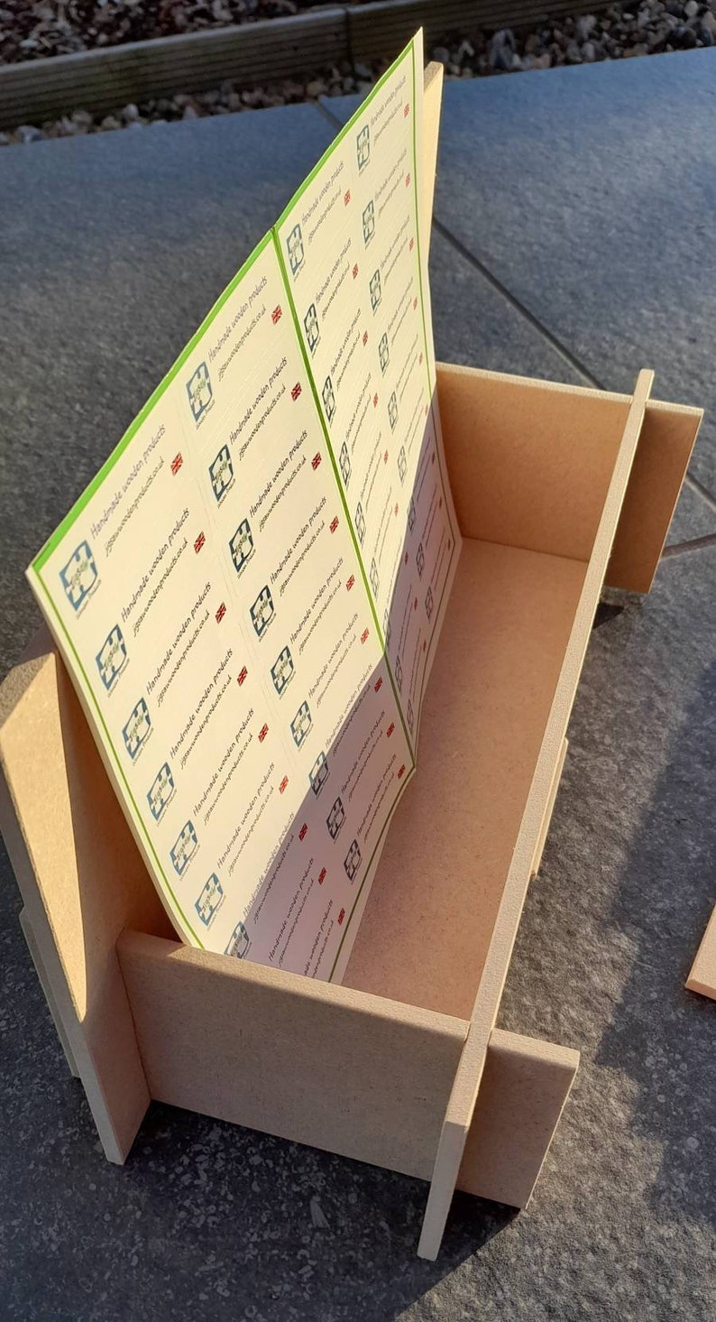May include: A light brown wooden box with a slotted design, holding a stack of printed labels. The labels have a green border and feature a logo and text that reads "Handmade wooden products". The box is sitting on a gray surface.