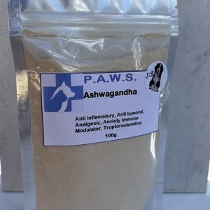 May include: A silver foil stand-up pouch containing 100g of beige powder. The label features a blue medical cross with a dog silhouette, the text P.A.W.S. Ashwagandha, a small dog sticker, and a list of properties including anti-inflammatory and analgesic.