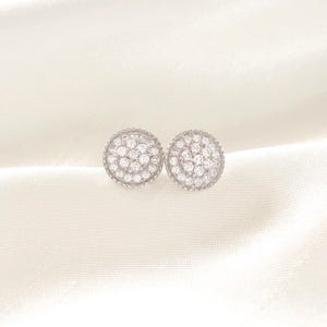 May include: A pair of silver-toned stud earrings. Each earring is round and covered in small, clear gemstones. The gemstones are arranged in concentric circles, creating a sparkling effect. The earrings are set against a white, textured background.