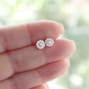 Op de afbeelding: Een paar gouden oorbel studs met een kleine parel in het midden en een halo van heldere zirkonia stenen.