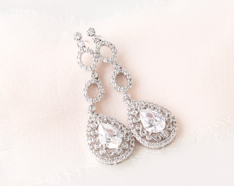 May include: A pair of silver-toned drop earrings featuring two circular links connected to a pear-shaped pendant. Each piece is encrusted with small clear stones and a larger central pear-cut clear stone, creating a sparkling, detailed design.