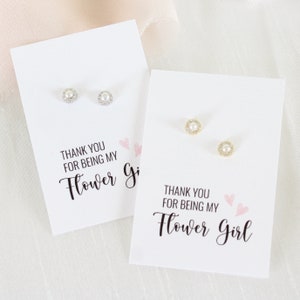 Op de afbeelding: Twee witte kaarten met de tekst "Bedankt dat je mijn bruidsmeisje bent" en twee paar parel oorbellen op elke kaart. De oorbellen zijn op witte kaarten met roze harten.