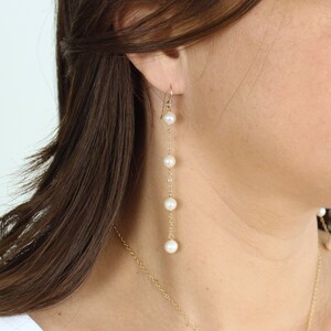 May include: A pair of long, gold chain earrings with white pearls. The earrings are dangling from the earlobe and have a total of five pearls.