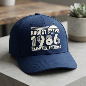 May include: A navy blue baseball cap featuring a cream-colored graphic print on the front. The design includes the text "AUGUST", "EST", "1986", and "LIMITED EDITION" alongside a stylized sun and mountain landscape graphic in a retro style.