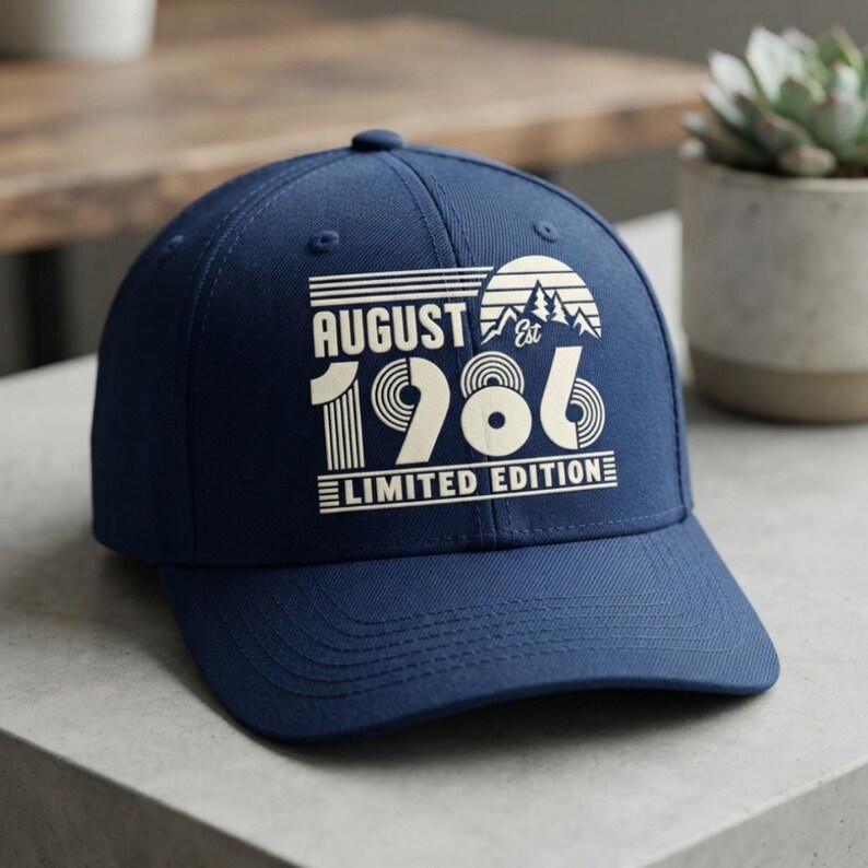 May include: A navy blue baseball cap featuring a cream-colored graphic print on the front. The design includes the text "AUGUST", "EST", "1986", and "LIMITED EDITION" alongside a stylized sun and mountain landscape graphic in a retro style.