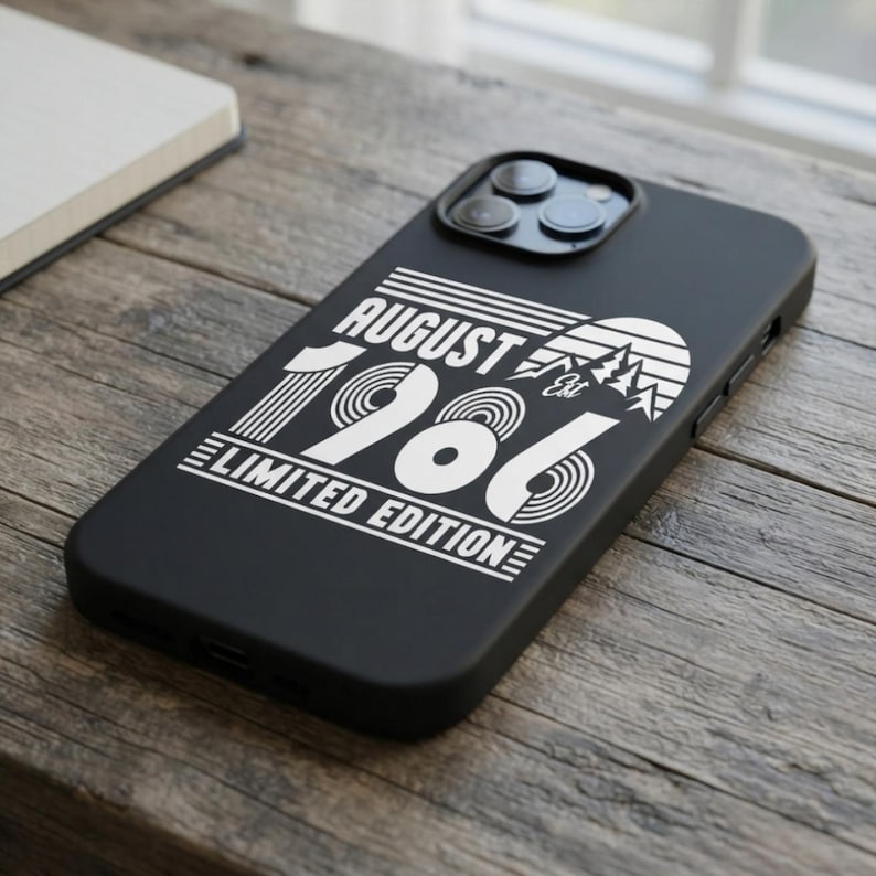 May include: A black smartphone case featuring a white graphic design with the text AUGUST 1986 LIMITED EDITION. The design includes a stylized mountain range, horizontal lines, and circular patterns, with the letters Est positioned near the peaks.