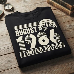 May include: A black crewneck sweatshirt featuring a cream-colored graphic print. The design includes the text AUGUST 1986, the word Est, and the phrase LIMITED EDITION. A stylized sun and mountain range graphic sits above the year 1986.