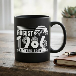 May include: A matte black ceramic mug featuring white printed text and graphics. The design includes the words AUGUST EST 1986 LIMITED EDITION, accompanied by a stylized mountain range and sun graphic with horizontal line accents.